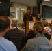 A military officer in dress uniform is delivering a speech at a podium bearing the 'Radboudumc' logo, likely at Radboud University Medical Center in the Netherlands. An audience of civilians and at least one other military personnel are seated facing the speaker. The setting appears to be a modern conference or lecture room.