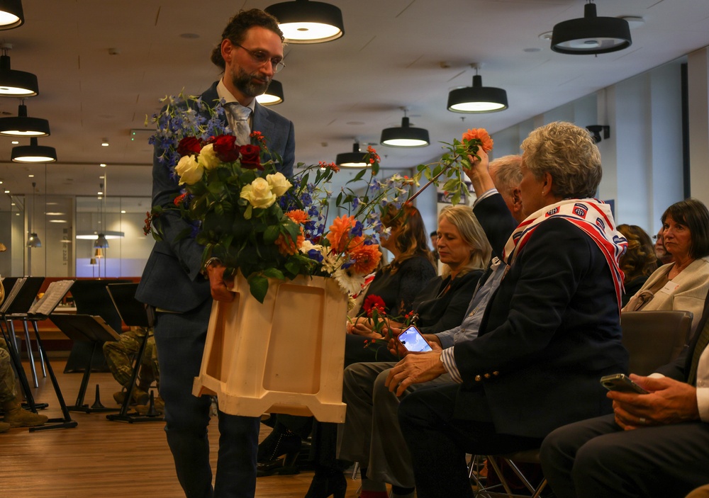 A formal indoor ceremony or event where a man in a blue suit is distributing flowers from a wooden tray to seated audience members. One seated person wearing a scarf with what appears to be a military unit insignia (82nd Airborne Division patch visible) is reaching up to take a flower. The setting appears to be a modern conference or event hall with pendant lighting and music stands visible in the background.