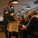 A formal indoor ceremony or event where a man in a blue suit is distributing flowers from a wooden tray to seated audience members. One seated person wearing a scarf with what appears to be a military unit insignia (82nd Airborne Division patch visible) is reaching up to take a flower. The setting appears to be a modern conference or event hall with pendant lighting and music stands visible in the background.