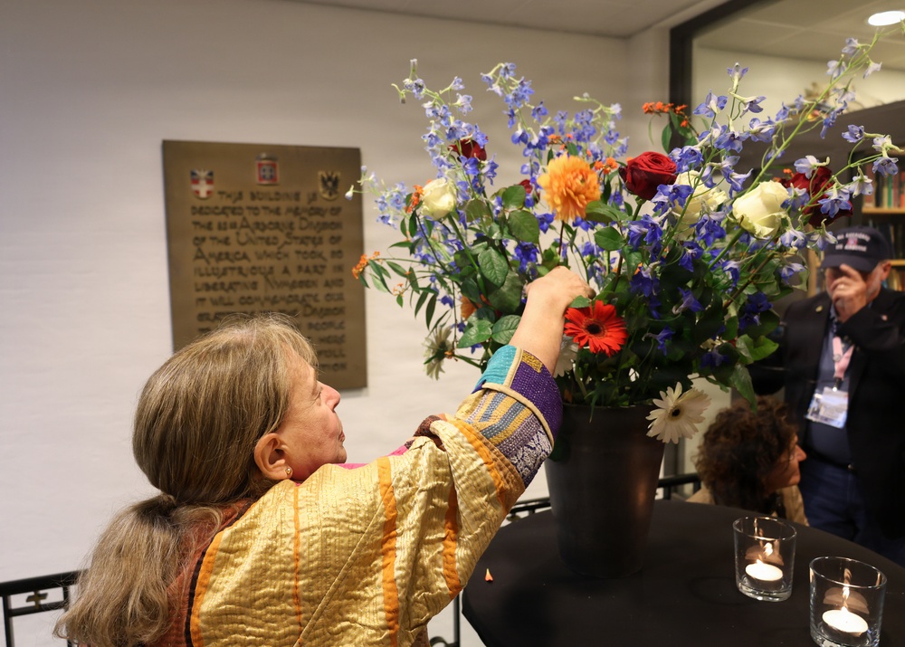 A woman in a colorful golden jacket arranges a large floral bouquet containing red roses, blue delphiniums, orange dahlias, gerbera daisies, and white flowers in a dark vase. The setting appears to be a memorial or commemorative venue, as evidenced by a bronze dedication plaque on the wall behind her referencing the U.S. Airborne Division and the liberation of Nijmegen. Candles in glass holders are visible on the table in the foreground.