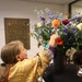 A woman in a colorful golden jacket arranges a large floral bouquet containing red roses, blue delphiniums, orange dahlias, gerbera daisies, and white flowers in a dark vase. The setting appears to be a memorial or commemorative venue, as evidenced by a bronze dedication plaque on the wall behind her referencing the U.S. Airborne Division and the liberation of Nijmegen. Candles in glass holders are visible on the table in the foreground.