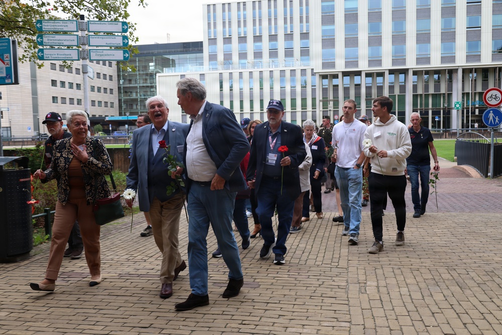 A group of people, mostly older adults, are walking together along a brick-paved path near a modern institutional building. Several participants are carrying flowers, including red carnations and white daisies, suggesting a commemorative or ceremonial procession. Directional signage in Dutch is visible in the background, indicating this is likely in the Netherlands.