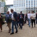 A group of people, mostly older adults, are walking together along a brick-paved path near a modern institutional building. Several participants are carrying flowers, including red carnations and white daisies, suggesting a commemorative or ceremonial procession. Directional signage in Dutch is visible in the background, indicating this is likely in the Netherlands.