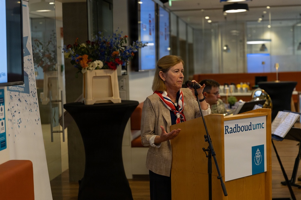 A woman is speaking at a podium bearing the 'Radboumc' logo, suggesting an official event at Radboud University Medical Center in the Netherlands. She is holding a microphone and gesturing with her hand, dressed in a blazer with a red, white, and blue scarf. The setting appears to be a modern indoor venue decorated with colorful flowers and featuring a small ensemble musician in the background.
