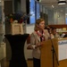 A woman is speaking at a podium bearing the 'Radboumc' logo, suggesting an official event at Radboud University Medical Center in the Netherlands. She is holding a microphone and gesturing with her hand, dressed in a blazer with a red, white, and blue scarf. The setting appears to be a modern indoor venue decorated with colorful flowers and featuring a small ensemble musician in the background.