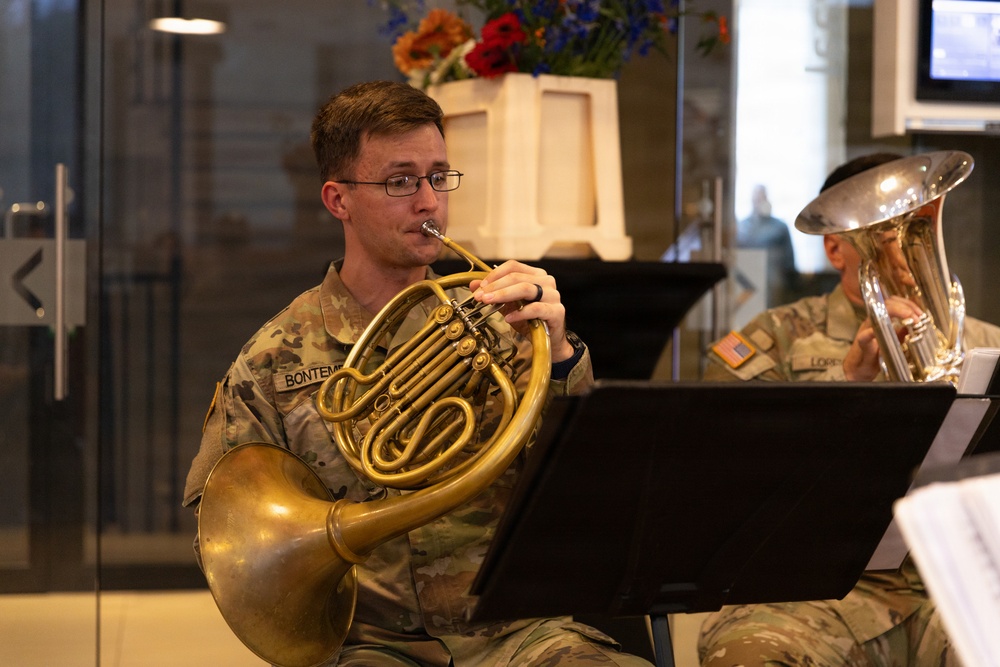 Two U.S. Army soldiers in MultiCam (OCP) uniforms are performing as musicians, reading sheet music from stands. The foreground soldier, identified by name tape as 'BONTEMPI', is playing a French horn, while the background soldier (partially visible, name tape reading 'LORE...') plays what appears to be a euphonium or tuba. The setting appears to be an indoor venue decorated with flowers, suggesting a formal or ceremonial performance.
