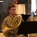 Two U.S. Army soldiers in MultiCam (OCP) uniforms are performing as musicians, reading sheet music from stands. The foreground soldier, identified by name tape as 'BONTEMPI', is playing a French horn, while the background soldier (partially visible, name tape reading 'LORE...') plays what appears to be a euphonium or tuba. The setting appears to be an indoor venue decorated with flowers, suggesting a formal or ceremonial performance.
