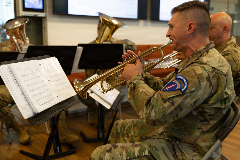 Military musicians in U.S. Army camouflage uniforms perform as a brass ensemble, reading from sheet music on stands. The foreground soldier plays a gold/brass trumpet while others behind him play larger brass instruments including what appear to be tubas or euphoniums. The patch on the foreground soldier's shoulder reads 'USAREUR AF BAND', identifying this as the United States Army Europe and Africa Band.