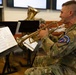 Military musicians in U.S. Army camouflage uniforms perform as a brass ensemble, reading from sheet music on stands. The foreground soldier plays a gold/brass trumpet while others behind him play larger brass instruments including what appear to be tubas or euphoniums. The patch on the foreground soldier's shoulder reads 'USAREUR AF BAND', identifying this as the United States Army Europe and Africa Band.