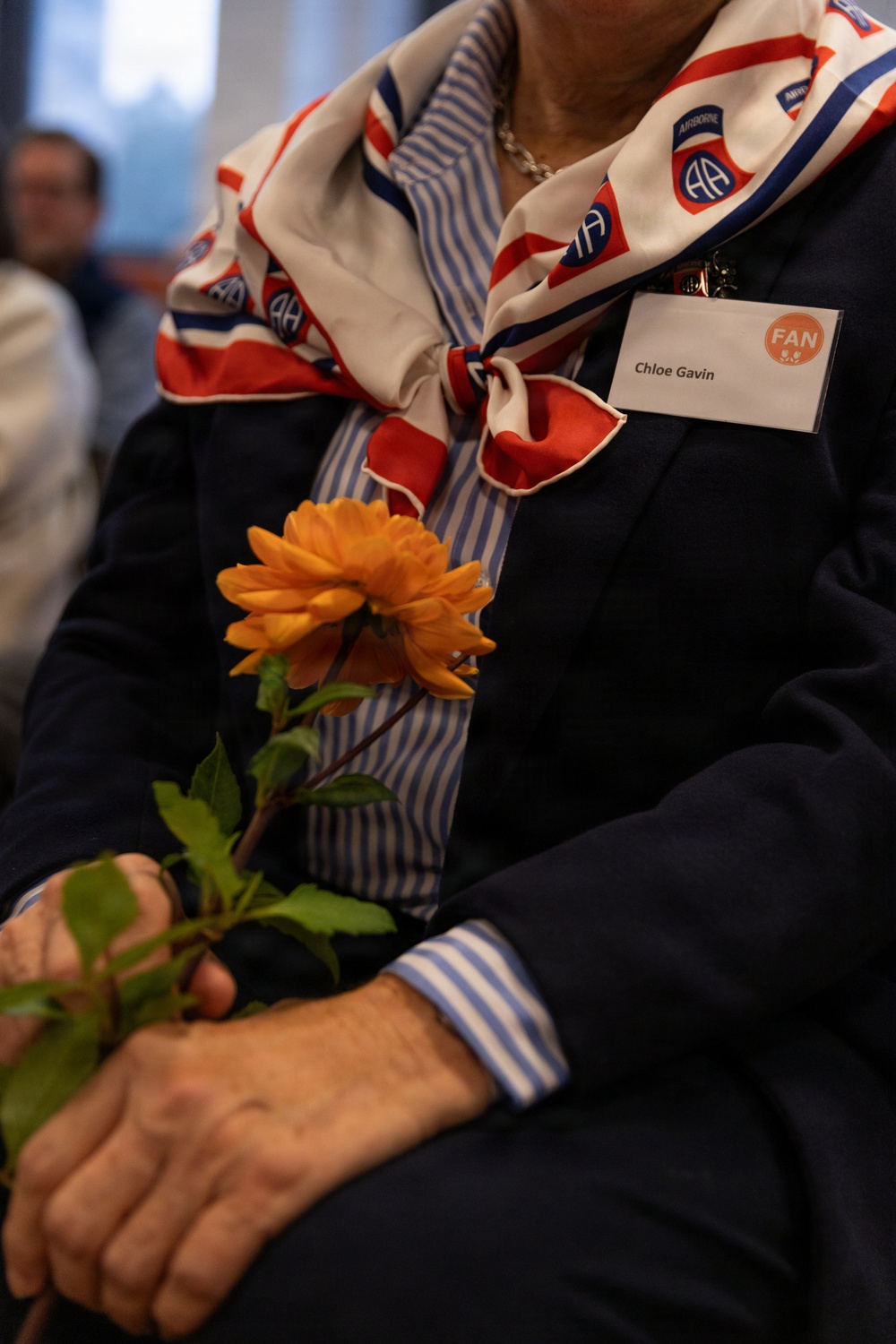 A person wearing a navy blazer and blue-striped shirt holds a single orange dahlia flower. They are wearing a red, white, and blue scarf featuring the 82nd Airborne Division 'AA' insignia, along with a name tag reading 'Chloe Gavin' with a 'FAN' designation. The setting appears to be an indoor event or ceremony.