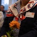 A person wearing a navy blazer and blue-striped shirt holds a single orange dahlia flower. They are wearing a red, white, and blue scarf featuring the 82nd Airborne Division 'AA' insignia, along with a name tag reading 'Chloe Gavin' with a 'FAN' designation. The setting appears to be an indoor event or ceremony.