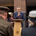 A man in a navy blue suit with a red tie is speaking at a wooden podium branded with the 'TALIS' logo, holding a microphone. The event appears to be a formal or commemorative ceremony, evidenced by the presence of a person in military uniform in the foreground and a person wearing a religious head covering (kippah). A gathered crowd of attendees is visible in the background near a brick building.