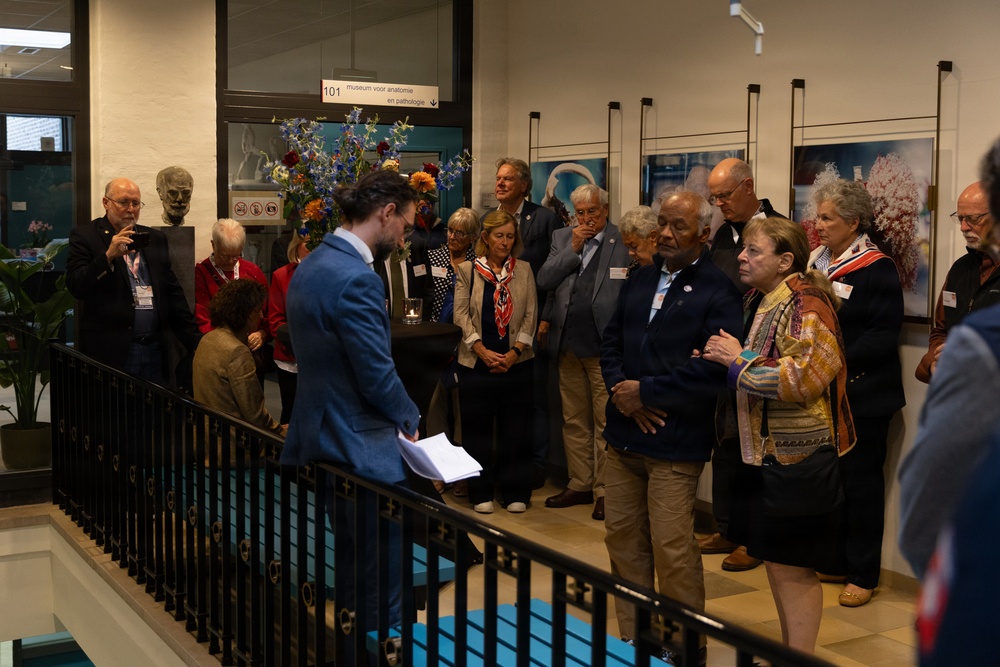 A group of approximately 20 adults are gathered in what appears to be the entrance or lobby of a museum for anatomy and pathology ('museum voor anatomie en pathologie'), as indicated by the sign numbered 101. A young man in a blue blazer appears to be addressing the group while holding papers, suggesting a guided tour introduction or formal presentation. The setting includes display photographs on the walls, floral arrangements, and a sculptural bust near the entrance.