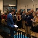 A group of approximately 20 adults are gathered in what appears to be the entrance or lobby of a museum for anatomy and pathology ('museum voor anatomie en pathologie'), as indicated by the sign numbered 101. A young man in a blue blazer appears to be addressing the group while holding papers, suggesting a guided tour introduction or formal presentation. The setting includes display photographs on the walls, floral arrangements, and a sculptural bust near the entrance.