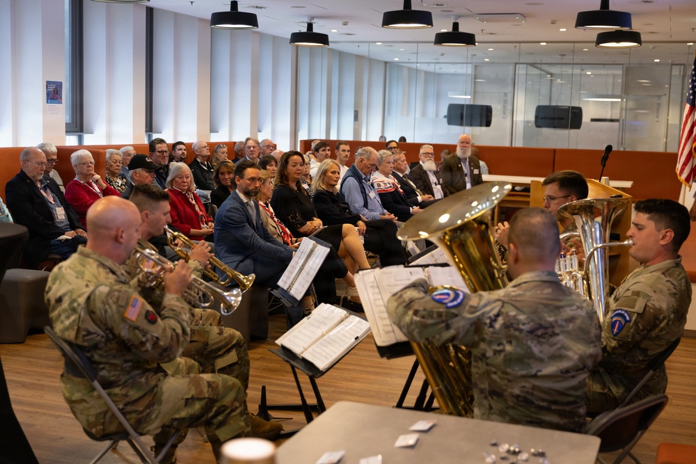 A military brass ensemble in U.S. Army camouflage uniforms performs for a seated audience in a modern indoor venue. The event appears to be a formal ceremony or commemorative gathering, with attendees wearing business attire and name badges. An American flag is visible in the background near a podium.