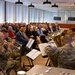 A military brass ensemble in U.S. Army camouflage uniforms performs for a seated audience in a modern indoor venue. The event appears to be a formal ceremony or commemorative gathering, with attendees wearing business attire and name badges. An American flag is visible in the background near a podium.