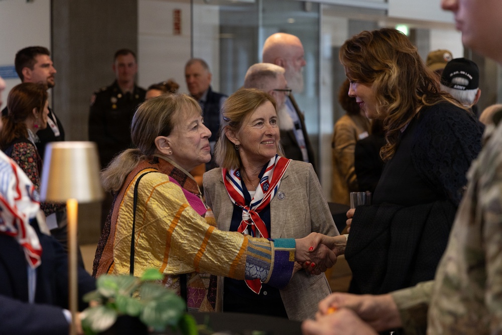 A formal reception or commemorative event is taking place in what appears to be a modern building lobby or conference center. Two women in the foreground are engaged in a handshake greeting, while military personnel and civilians mingle in the background. The presence of military uniforms, a patriotic scarf, and a veteran's cap suggests this may be a military or veterans-related ceremony or gathering.