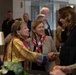 A formal reception or commemorative event is taking place in what appears to be a modern building lobby or conference center. Two women in the foreground are engaged in a handshake greeting, while military personnel and civilians mingle in the background. The presence of military uniforms, a patriotic scarf, and a veteran's cap suggests this may be a military or veterans-related ceremony or gathering.