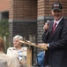 An older man in a dark suit and red tie is speaking at an outdoor podium while holding a microphone. He is wearing a black military-style cap with what appears to be an 82nd Airborne Division insignia. An audience of mostly older adults is seated and listening in the background.