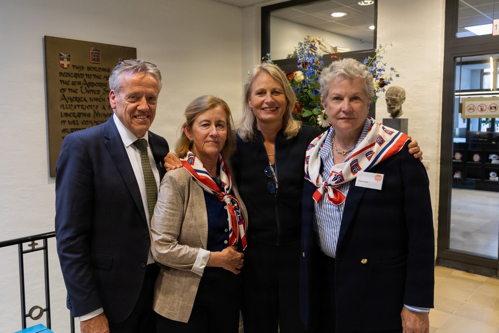Four individuals pose for a group photo inside what appears to be a museum or memorial building dedicated to the 82nd Airborne Division of the United States Army. Three of the four individuals are wearing red, white, and blue scarves bearing the 82nd Airborne Division insignia. A commemorative plaque referencing the 82nd Airborne Division and the liberation of Nijmegen is visible on the wall behind them.