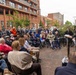 An outdoor public ceremony or commemoration event is taking place in an urban plaza surrounded by modern red-brick buildings. A woman is speaking at a podium with a microphone to a seated audience of approximately 40-50 people, including elderly individuals, some in wheelchairs or with mobility aids, and several people in military uniforms. An American flag and what appears to be another national flag are visible in the background.