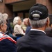An outdoor gathering of predominantly elderly individuals, likely a veterans' commemoration or memorial event. A man in the foreground wears a black cap embroidered with 'IN AIR, ON LAND' and 'ARMY', indicating U.S. Army service. A woman beside him wears a red, white, and blue scarf bearing the 82nd Airborne Division patch.