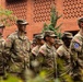 A formation of U.S. Army soldiers standing at attention in military dress uniforms (OCP/MultiCam camouflage pattern). Several soldiers display the 10th Mountain Division patch on their left shoulders, and all wear the U.S. flag patch on their right shoulders. The formation appears to be taking place outdoors in front of a red brick building, likely during a formal ceremony or parade.