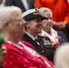 A female naval officer in dress uniform is seated among civilians at what appears to be a formal outdoor ceremony or event. She is smiling and looking forward, wearing a dark service dress uniform with multiple ribbons and a white-topped officer's cap with gold insignia. The surrounding attendees are dressed in civilian clothing, suggesting a public or commemorative event.