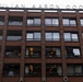 The image shows the facade of a multi-story brick building with the name 'JULIAN AARON COOK' displayed in large letters along the roofline. The building features a modernist architectural style with large rectangular windows, balconies with plants and furniture, and vertical brick pilasters. The lower portion of the building also displays partial lettering spelling 'AARON'.