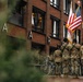 A military color guard in U.S. Army camouflage uniforms stands at attention carrying the American flag and what appears to be a unit guidon during an outdoor ceremony. The formation is positioned in front of a modern red-brick building in what appears to be an urban setting. A Ukrainian flag is also visible in the background near a seated audience.