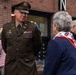 A U.S. Army officer in dress uniform, identified by name tag as 'Wright', converses with an elderly woman wearing a scarf bearing the 82nd Airborne Division patch insignia. The setting appears to be an outdoor commemorative or ceremonial event, likely related to the 82nd Airborne Division given the matching insignia visible on both the officer's shoulder patch and the woman's scarf. Additional military personnel in modern combat uniforms are visible in the background.