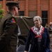 A military officer in olive drab dress uniform is engaged in conversation with a civilian woman wearing a navy blazer and a red, white, and blue scarf. The setting appears to be an outdoor ceremonial or commemorative event, with a vintage military vehicle visible in the background. Several other attendees, including additional military personnel in modern camouflage uniforms, are present in the background.