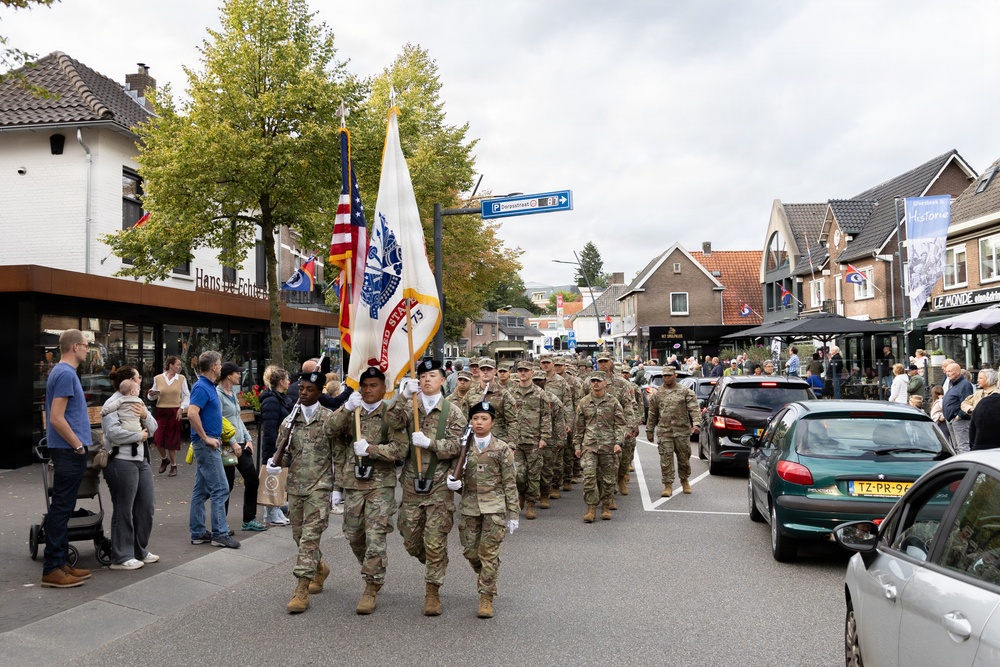 A column of U.S. Army soldiers in camouflage uniforms marches through a Dutch town street, led by a color guard carrying the American flag and a U.S. Army unit guidon. Civilians line the sidewalks watching the parade, and Dutch street signs, license plates, and architecture confirm the location as the Netherlands. The event appears to be a commemorative military parade, likely related to WWII liberation anniversaries.