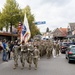 A column of U.S. Army soldiers in camouflage uniforms marches through a Dutch town street, led by a color guard carrying the American flag and a U.S. Army unit guidon. Civilians line the sidewalks watching the parade, and Dutch street signs, license plates, and architecture confirm the location as the Netherlands. The event appears to be a commemorative military parade, likely related to WWII liberation anniversaries.