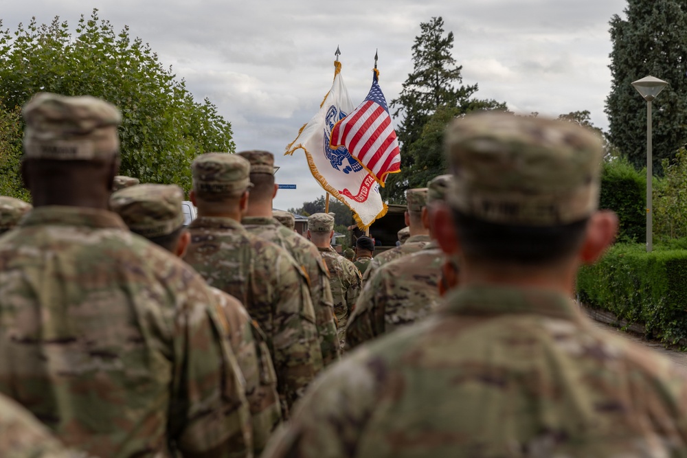 A formation of U.S. Army soldiers in Operational Camouflage Pattern (OCP) uniforms stands at attention during what appears to be a military ceremony or parade. In the background, a color guard carries the U.S. national flag and a U.S. Army organizational flag with gold fringe. The setting appears to be an outdoor military installation, likely in Europe based on the surrounding architecture and vegetation.