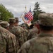 A formation of U.S. Army soldiers in Operational Camouflage Pattern (OCP) uniforms stands at attention during what appears to be a military ceremony or parade. In the background, a color guard carries the U.S. national flag and a U.S. Army organizational flag with gold fringe. The setting appears to be an outdoor military installation, likely in Europe based on the surrounding architecture and vegetation.