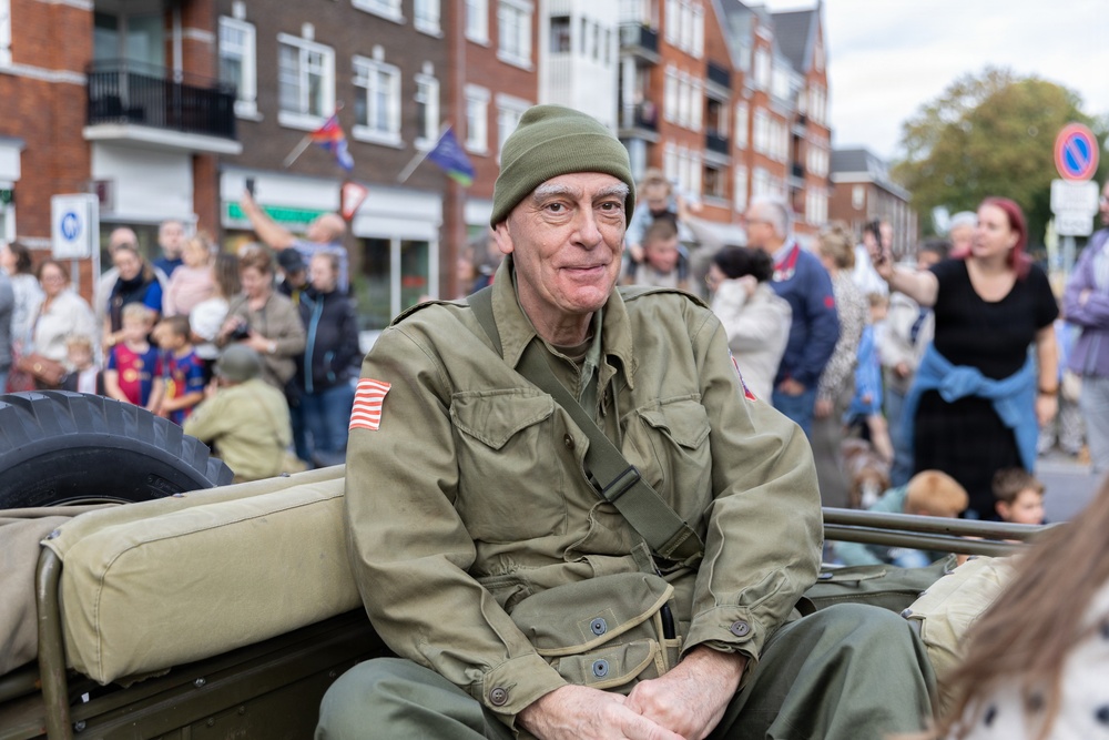 An older man wearing WWII-era U.S. Army uniform with an American flag patch on the sleeve sits in what appears to be a vintage military jeep during a public parade or commemoration event. He is wearing an olive drab field jacket and knit cap, smiling at the camera. A crowd of spectators lines the street behind him in an urban setting with brick buildings.
