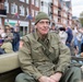 An older man wearing WWII-era U.S. Army uniform with an American flag patch on the sleeve sits in what appears to be a vintage military jeep during a public parade or commemoration event. He is wearing an olive drab field jacket and knit cap, smiling at the camera. A crowd of spectators lines the street behind him in an urban setting with brick buildings.