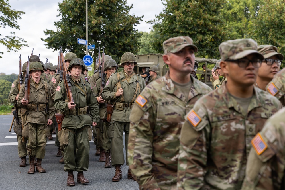 The image shows a military parade or commemorative march featuring two distinct groups of soldiers. On the left, participants are dressed in World War II-era U.S. Army uniforms with M1 helmets and carrying period-correct rifles (M1 Garands). On the right, modern U.S. Army soldiers in contemporary MultiCam/OCP camouflage uniforms observe or participate alongside them, suggesting a memorial or anniversary commemoration event.