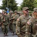 The image shows a military parade or commemorative march featuring two distinct groups of soldiers. On the left, participants are dressed in World War II-era U.S. Army uniforms with M1 helmets and carrying period-correct rifles (M1 Garands). On the right, modern U.S. Army soldiers in contemporary MultiCam/OCP camouflage uniforms observe or participate alongside them, suggesting a memorial or anniversary commemoration event.