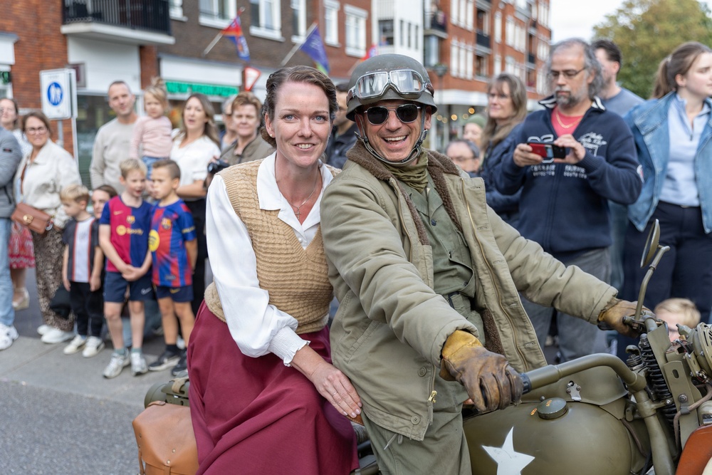 A historical reenactment or commemorative parade is taking place on a town street, featuring two participants dressed in WWII-era costumes riding an olive-drab military motorcycle with a white star insignia. A woman in 1940s civilian attire sits behind a man dressed as an Allied soldier wearing a period helmet, goggles, and military jacket. A crowd of modern-day spectators lines the street in the background.