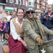 A historical reenactment or commemorative parade is taking place on a town street, featuring two participants dressed in WWII-era costumes riding an olive-drab military motorcycle with a white star insignia. A woman in 1940s civilian attire sits behind a man dressed as an Allied soldier wearing a period helmet, goggles, and military jacket. A crowd of modern-day spectators lines the street in the background.