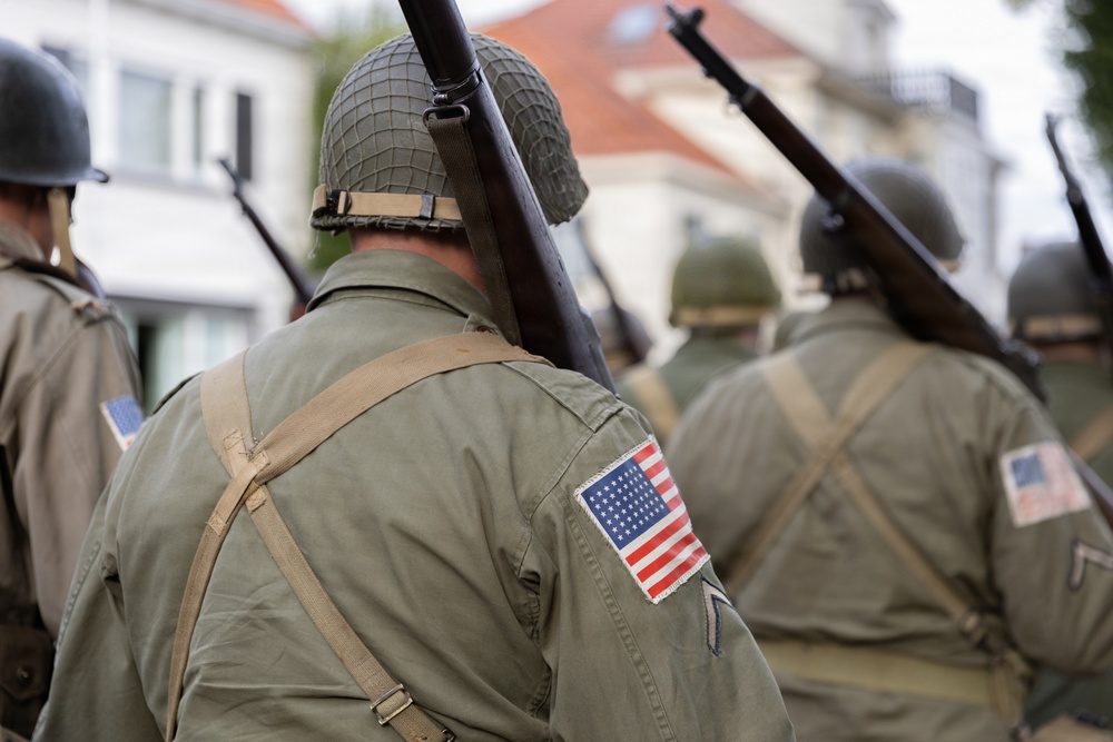 The image shows a group of individuals dressed in WWII-era U.S. Army uniforms marching in what appears to be a historical reenactment or commemorative parade. They are wearing M1 steel helmets with netting, olive drab field jackets bearing American flag patches, and carrying period-correct rifles such as the M1 Garand. The setting appears to be a European town street, consistent with liberation commemoration events.