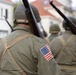 The image shows a group of individuals dressed in WWII-era U.S. Army uniforms marching in what appears to be a historical reenactment or commemorative parade. They are wearing M1 steel helmets with netting, olive drab field jackets bearing American flag patches, and carrying period-correct rifles such as the M1 Garand. The setting appears to be a European town street, consistent with liberation commemoration events.