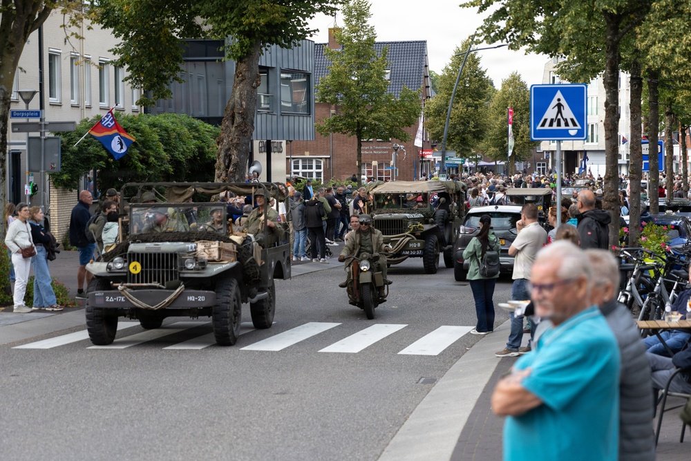 A military vehicle parade is taking place on a town street, featuring vintage WWII-era jeeps, trucks, and a motorcycle with riders dressed in period military uniforms. Large crowds of spectators line the sidewalks to watch the procession. The setting appears to be a Dutch town, as indicated by the street sign reading 'Dorpsplein' and Dutch license plates on the vehicles.
