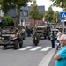 A military vehicle parade is taking place on a town street, featuring vintage WWII-era jeeps, trucks, and a motorcycle with riders dressed in period military uniforms. Large crowds of spectators line the sidewalks to watch the procession. The setting appears to be a Dutch town, as indicated by the street sign reading 'Dorpsplein' and Dutch license plates on the vehicles.