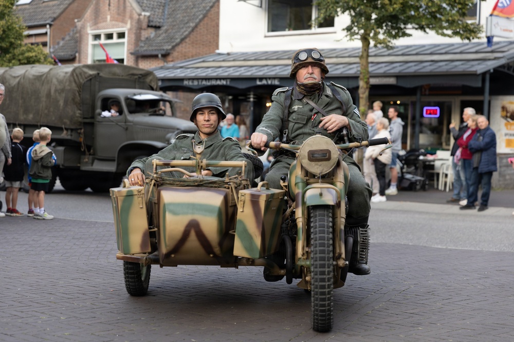 Two individuals dressed in WWII-era German military uniforms ride a camouflage-painted motorcycle with sidecar through what appears to be a town square or street. A vintage military truck is visible in the background, suggesting this is a historical reenactment or commemorative parade event. Civilian spectators line the street and a restaurant called 'La Vie' is visible in the background.