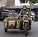 Two individuals dressed in WWII-era German military uniforms ride a camouflage-painted motorcycle with sidecar through what appears to be a town square or street. A vintage military truck is visible in the background, suggesting this is a historical reenactment or commemorative parade event. Civilian spectators line the street and a restaurant called 'La Vie' is visible in the background.