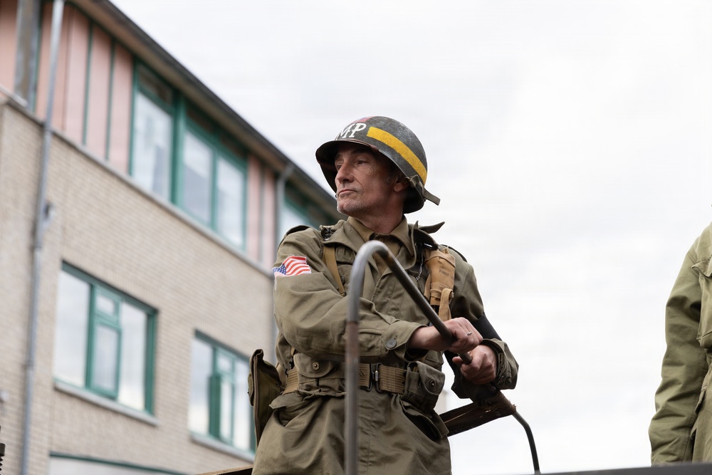 A man dressed in WWII-era U.S. Army Military Police (MP) uniform stands atop what appears to be a military vehicle. He wears an M1 helmet marked 'MP' with a yellow stripe, an olive drab field jacket with a U.S. flag patch, and carries military equipment. The setting appears to be a modern street with a brick building in the background, suggesting a historical reenactment or commemorative event.