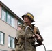A man dressed in WWII-era U.S. Army Military Police (MP) uniform stands atop what appears to be a military vehicle. He wears an M1 helmet marked 'MP' with a yellow stripe, an olive drab field jacket with a U.S. flag patch, and carries military equipment. The setting appears to be a modern street with a brick building in the background, suggesting a historical reenactment or commemorative event.